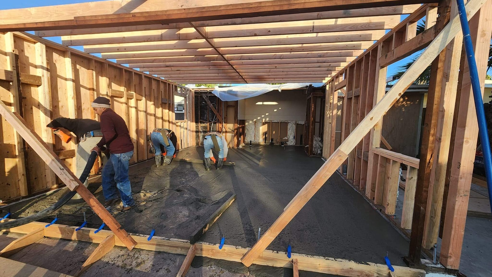 Crew finishing a fresh concrete slab inside a wood-framed garage conversion shell at dusk