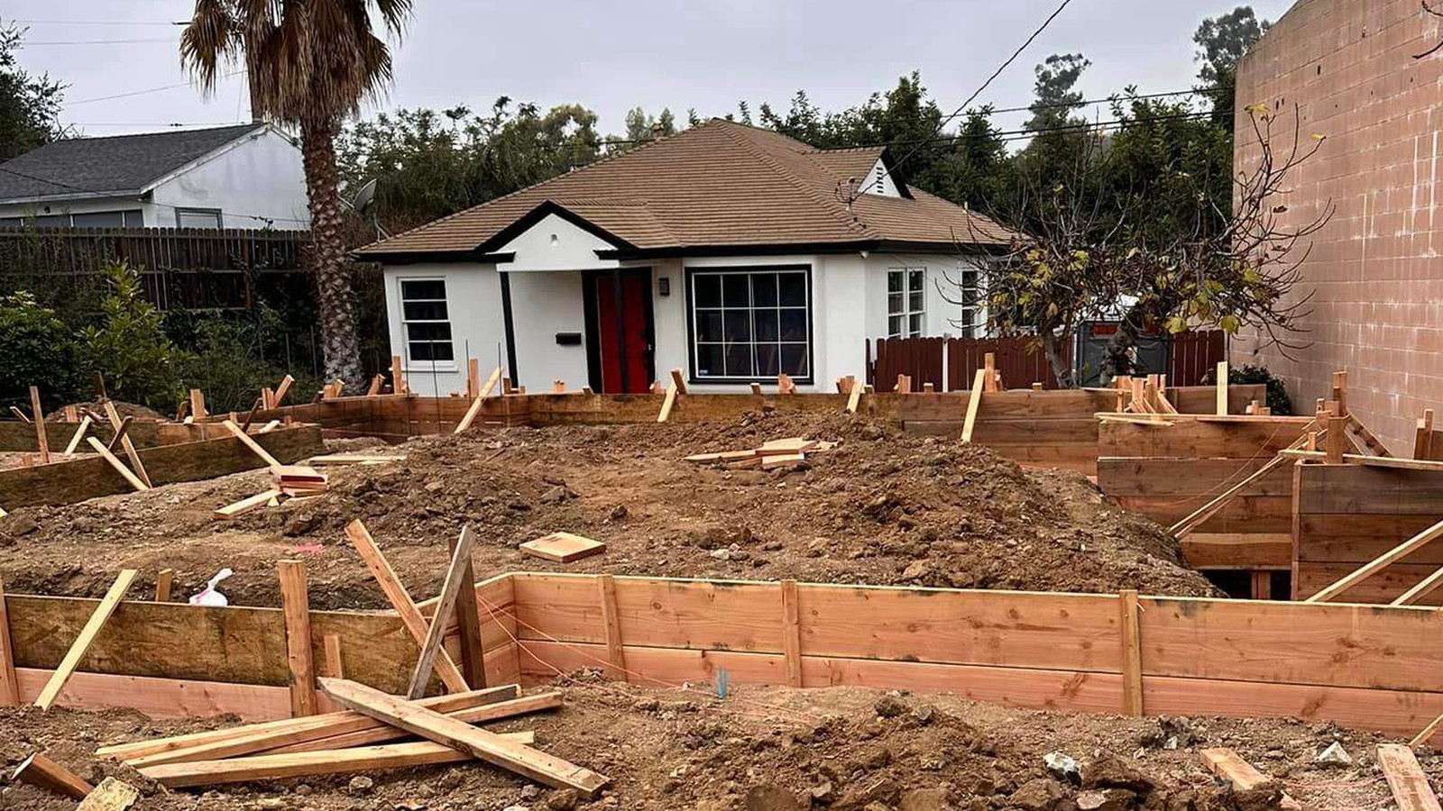 Foundation work underway at an older single-story house, with the ground opened up and redwood formwork staked across the excavation