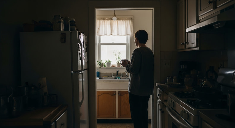A person stands in the doorway of an ordinary, slightly dated kitchen in the early morning, looking into the room with a mug in one hand.