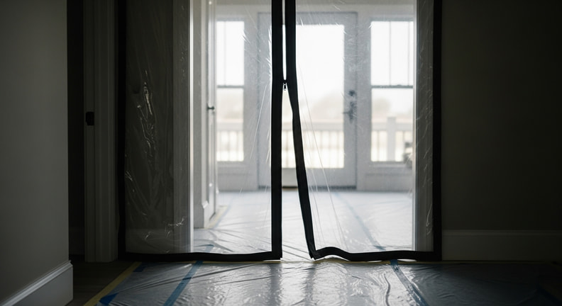 A heavy plastic dust barrier with a zippered opening seals a doorway in a home, with floor protection running up to it and daylight visible through the sheeting.