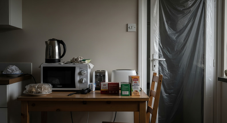A family kitchen table set up as a temporary cooking area with a kettle, a microwave and boxed provisions, with plastic sheeting visible in the doorway behind.