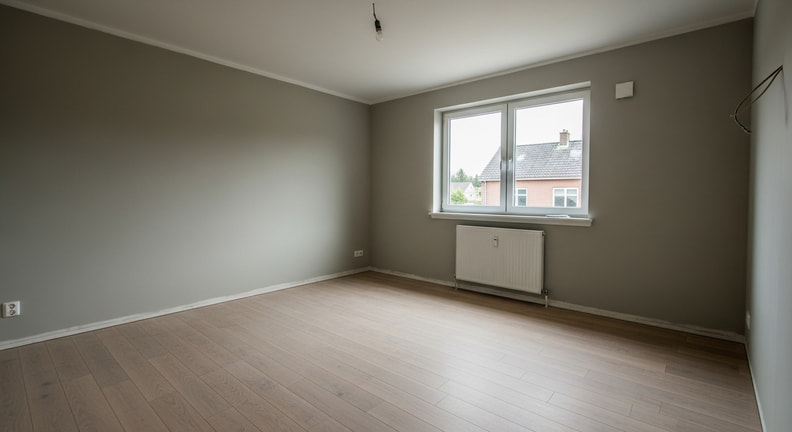 A finished, clean and empty room with new flooring and freshly painted walls, entirely free of tools, materials and protective covering, lit by daylight from a window.